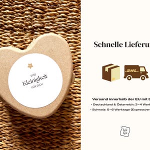 May include: A heart-shaped, light brown box with a white circular label. The label has the text "EINE Kleinigkeit F&Uuml;R DICH" in black. The box is on a woven, brown surface.