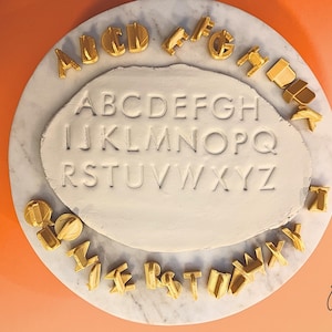 May include: A craft image shows a white clay oval with the alphabet pressed into it. Surrounding the clay are gold-colored letter-shaped cookie cutters. The clay and cutters sit on a round, marble-patterned surface against an orange background. The image is well-lit.