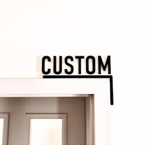 May include: A black sign with the word "CUSTOM" in all caps, mounted above a white door frame.