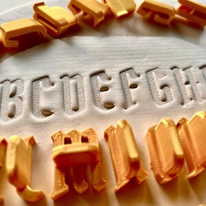 May include: A set of gold-colored plastic alphabet stamps arranged around a white circular clay surface. The letters A through I are clearly impressed into the clay, while the individual three-dimensional letter stamps are scattered around.