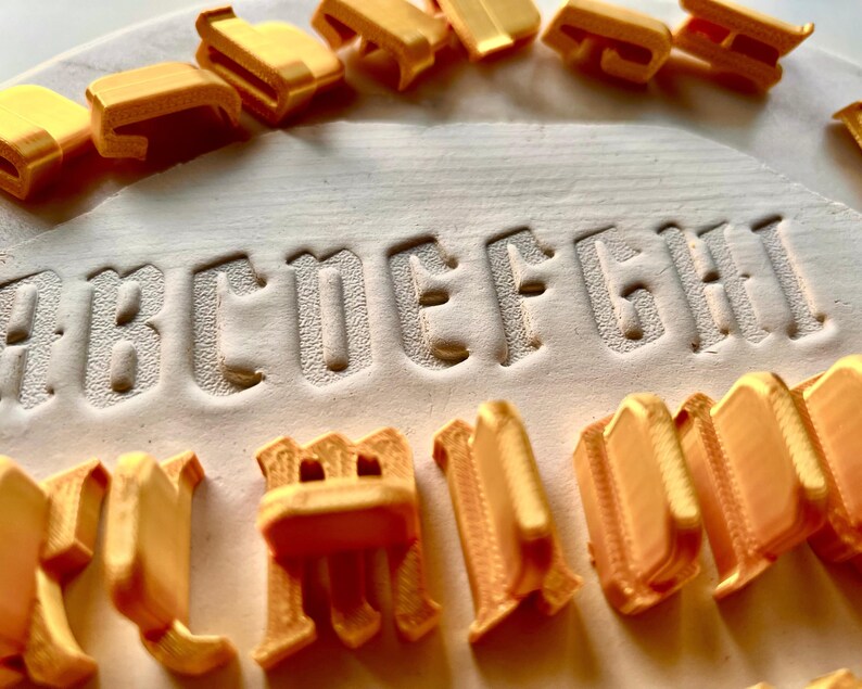 May include: A set of gold-colored plastic alphabet stamps arranged around a white circular clay surface. The letters A through I are clearly impressed into the clay, while the individual three-dimensional letter stamps are scattered around.