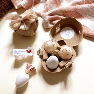 Pu&ograve; includere: Una composizione a tema pasquale. Un cartone per uova contiene tre uova macchiate. Una piccola scatola regalo rotonda &egrave; legata con un fiocco di iuta. Un contenitore bianco a forma di carota contiene denaro arrotolato. Una carta dice "Alles Gute zum 10. Schl&uuml;pftag!"