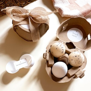 Könnte beinhalten: Eine herzförmige Geschenkbox mit einer Schleife aus Jute, ein Eierkarton aus Pappe mit drei gesprenkelten eiförmigen Kerzen und ein kleiner weißer Behälter mit einer Karte mit der Aufschrift "Du wirst Uroma". Der Eierkarton hat einen Aufkleber mit der Aufschrift "Knack mich!"