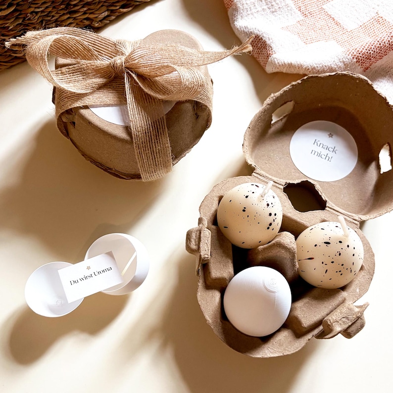 May include: A heart-shaped gift box tied with a burlap bow, a cardboard egg carton holding three speckled egg-shaped candles, and a small white container with a card that reads "Du wirst Uroma". The egg carton has a sticker that reads "Knack mich!"