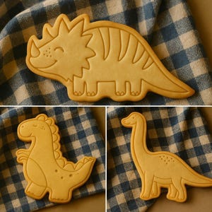 May include: Three dinosaur-shaped cookies on a blue and white checkered cloth. The cookies are light brown and feature different dinosaur designs: a triceratops, a stegosaurus, and a long-necked dinosaur. The cookies are detailed with etched lines.