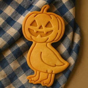 May include: A Halloween-themed cookie in the shape of a pumpkin-headed duck. The cookie is light orange and rests on a blue and white checkered cloth. The pumpkin face has carved-out eyes and a smiling mouth.