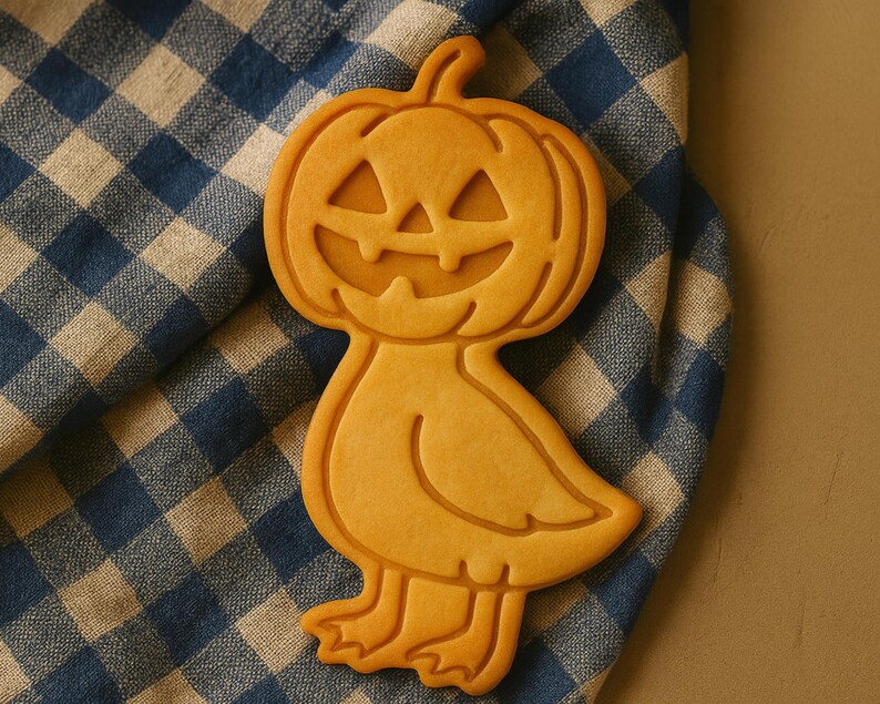 May include: A Halloween-themed cookie in the shape of a pumpkin-headed duck. The cookie is light orange and rests on a blue and white checkered cloth. The pumpkin face has carved-out eyes and a smiling mouth.