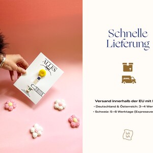 Op de afbeelding: Een houten wasknijper met een gele pompon aan de bovenkant, bevestigd op een witte kaart met de tekst ALLES GUTE. Vier kleine, gehaakte bloemvormige versieringen in roze en wit zijn rondom de kaart gerangschikt.