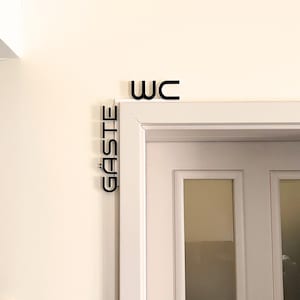 May include: Black WC and G&Auml;STE signs mounted above a white door. The word WC is above the door frame, and the word G&Auml;STE is vertically aligned on the left side of the door frame. The signs are in a sans-serif font.