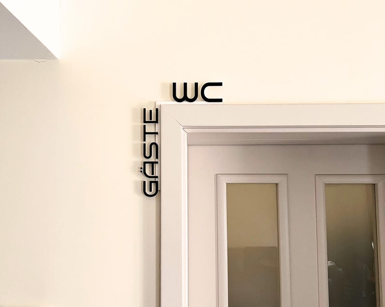 May include: Black WC and G&Auml;STE signs mounted above a white door. The word WC is above the door frame, and the word G&Auml;STE is vertically aligned on the left side of the door frame. The signs are in a sans-serif font.