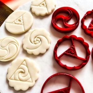 Könnte beinhalten: Ein Set roter Plastik-Ausstecher und einfache weiße Kekse mit verschiedenen geometrischen Mustern. Die Muster umfassen eine Spirale, ein Dreieck und ein Yin-Yang-Symbol.