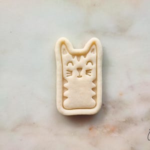 May include: A beige cat-shaped cookie cutter with a rectangular outline. The cat has a smiling face, whiskers, and a striped pattern. The cookie cutter is on a marble surface.