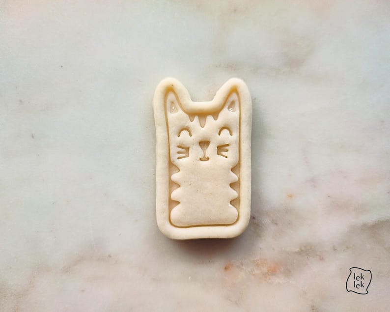 May include: A beige cat-shaped cookie cutter with a rectangular outline. The cat has a smiling face, whiskers, and a striped pattern. The cookie cutter is on a marble surface.