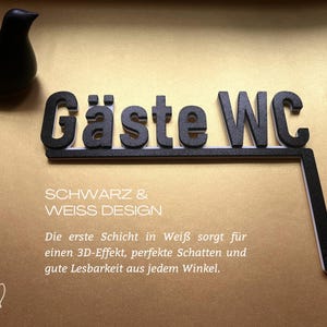 May include: Black "G&auml;ste WC" sign with white trim, and a black bird figurine on a gold background. The sign includes the text "SCHWARZ & WEISS DESIGN" and additional German text.