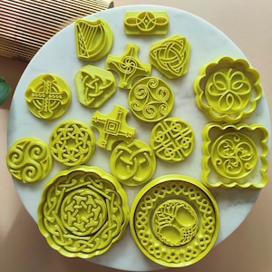 May include: A set of 14 yellow plastic cookie cutters featuring various Irish symbols, including shamrocks, Celtic knots, and crosses. The cutters are arranged on a white surface.