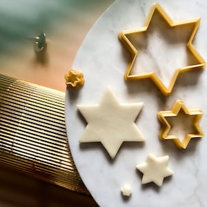 Op de afbeelding: Vijf gouden stervormige koekjesuitstekers in verschillende maten op een wit marmeren oppervlak. De grootste uitsteker is ongeveer 10 cm breed.