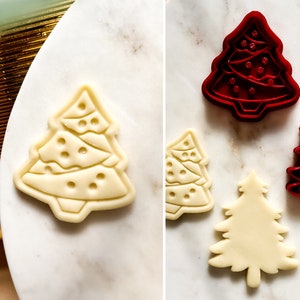 Puede incluir: Dos cortadores de galletas con forma de árbol de Navidad, uno rojo y otro blanco, sobre una superficie de mármol blanco. El cortador rojo está decorado con un patrón de adornos y luces. El cortador blanco es liso.