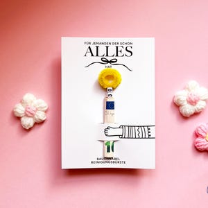 May include: A white rectangular card featuring the text F&Uuml;R JEMANDEN DER SCHON ALLES HAT, a yellow fuzzy ring, a rolled ten euro banknote, and the text BAUCHNABEL REINIGUNGSB&Uuml;RSTE. Two small crocheted flowers in white and pink flank the card.