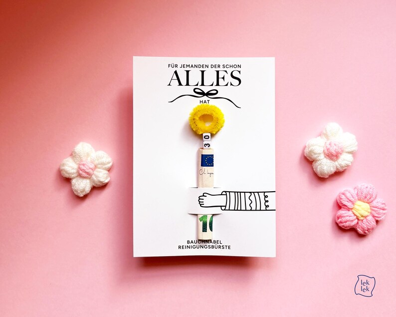 May include: A white rectangular card featuring the text F&Uuml;R JEMANDEN DER SCHON ALLES HAT, a yellow fuzzy ring, a rolled ten euro banknote, and the text BAUCHNABEL REINIGUNGSB&Uuml;RSTE. Two small crocheted flowers in white and pink flank the card.