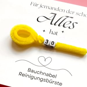 May include: A white card with German text and a yellow pipe cleaner shaped like a navel brush. The text reads "F&uuml;r jemanden der schon Alles hat" and "Bauchnabel Reinigungsb&uuml;rste". The number "30" is displayed on the brush.