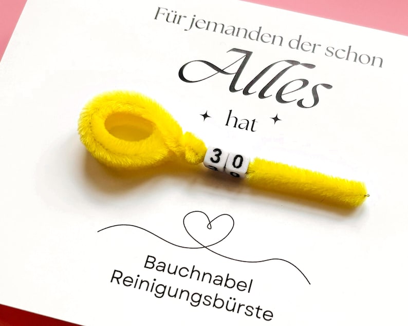May include: A white card with German text and a yellow pipe cleaner shaped like a navel brush. The text reads "F&uuml;r jemanden der schon Alles hat" and "Bauchnabel Reinigungsb&uuml;rste". The number "30" is displayed on the brush.