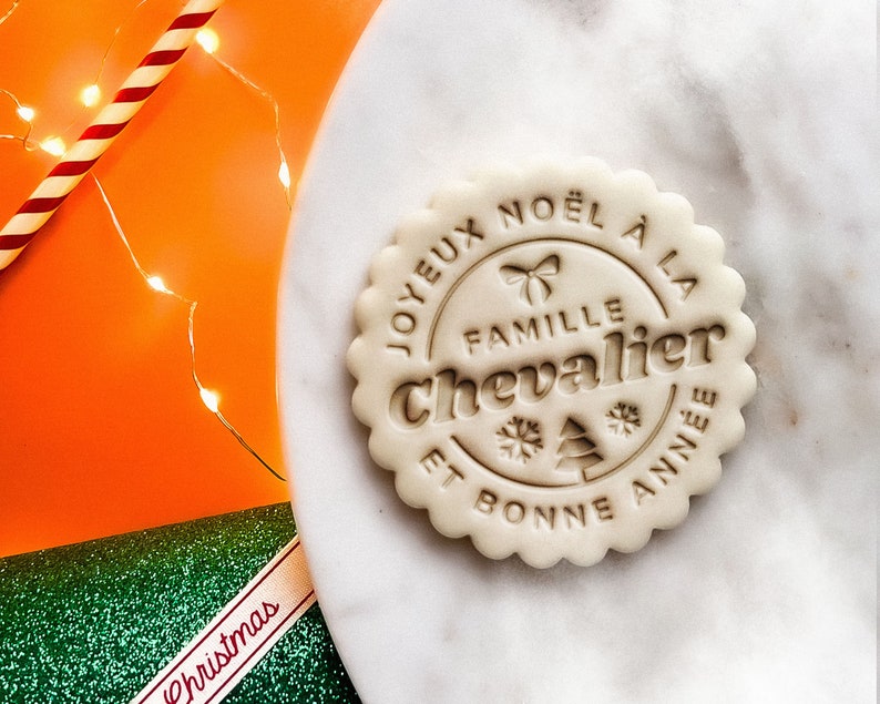 May include: A white cookie with a brown inscription that reads "Joyeux Noel a la Famille Chevalier et Bonne Annee" with a snowflake and a Christmas tree design.