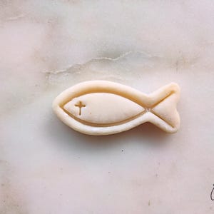 May include: A light beige fish-shaped object with a cross symbol carved into the center. The object is set against a marble background. The fish has a detailed outline and a tail fin.