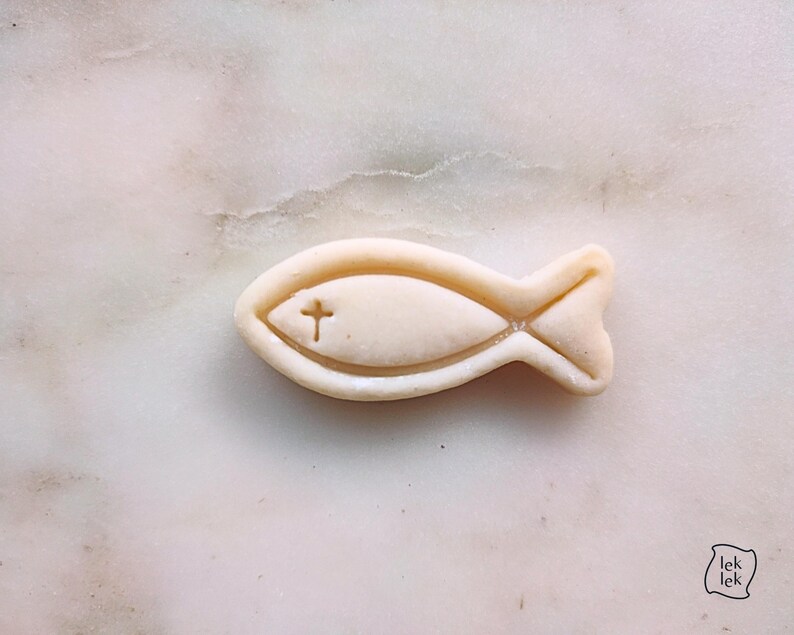 May include: A light beige fish-shaped object with a cross symbol carved into the center. The object is set against a marble background. The fish has a detailed outline and a tail fin.