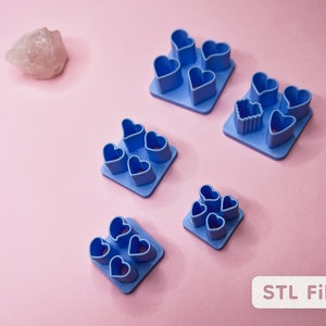 May include: Five different blue plastic heart-shaped cookie cutters, each with multiple heart-shaped molds. The cutters are arranged on a pink surface. STL Files.