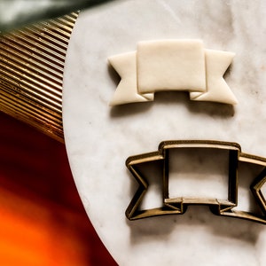 Op de afbeelding: Een gouden metalen koekjesvormpje in de vorm van een banner met een witte fondantbanner op een wit marmeren oppervlak.
