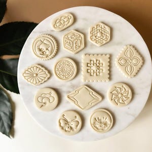 Puede incluir: Un juego de 12 cortadores de galletas con varios diseños geométricos y florales. Los cortadores están hechos de un material marrón claro y están dispuestos sobre una superficie de mármol blanco.