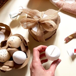 Pu&ograve; includere: Un assortimento di oggetti decorativi, tra cui candele a forma di uovo macchiato in un cartone per uova, una scatola a forma di cuore legata con un fiocco di juta e un oggetto rotondo bianco con una fessura. Il testo "Knack mich!" &egrave; visibile.