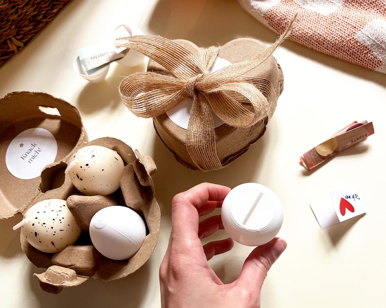 Pu&ograve; includere: Un assortimento di oggetti decorativi, tra cui candele a forma di uovo macchiato in un cartone per uova, una scatola a forma di cuore legata con un fiocco di juta e un oggetto rotondo bianco con una fessura. Il testo "Knack mich!" &egrave; visibile.