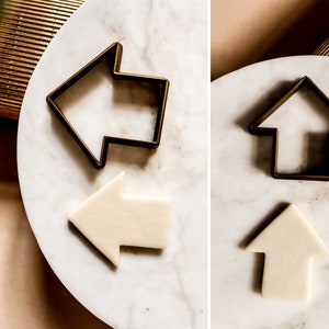 May include: Two gold metal arrow cookie cutters and two white fondant arrows on a white marble surface.