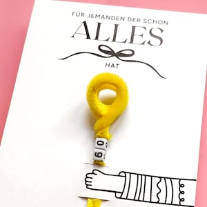 May include: White greeting card with the German text "F&Uuml;R JEMANDEN DER SCHON ALLES HAT" and a yellow fuzzy decoration. The decoration has the number "60" on it. A cartoon hand is also visible.