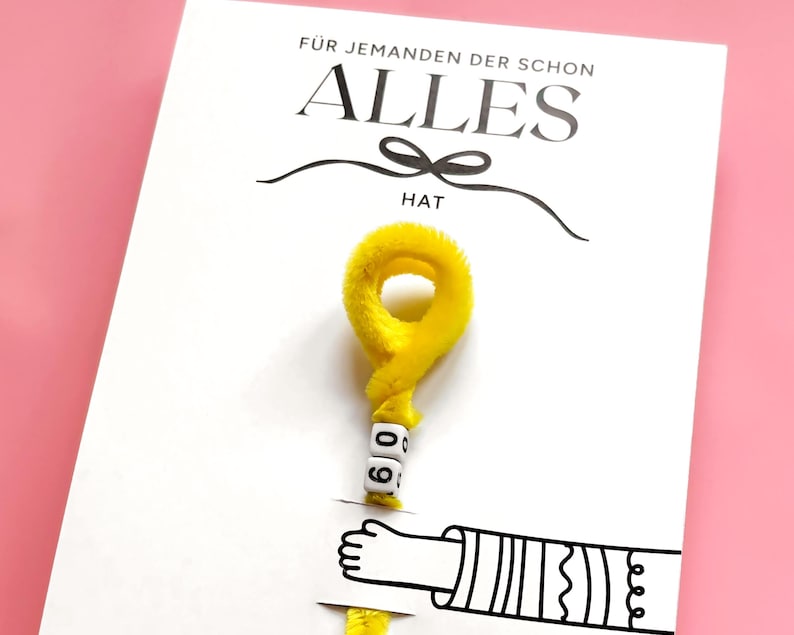 May include: White greeting card with the German text "F&Uuml;R JEMANDEN DER SCHON ALLES HAT" and a yellow fuzzy decoration. The decoration has the number "60" on it. A cartoon hand is also visible.