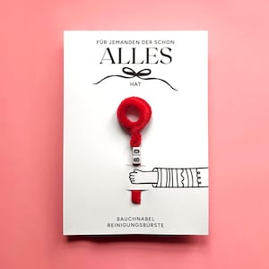 Peut inclure: Une carte blanche avec du texte allemand et un objet rouge duveteux. La carte indique "F&Uuml;R JEMANDEN DER SCHON ALLES HAT" et "BAUCHNABEL REINIGUNGSB&Uuml;RSTE". L'objet rouge est attach&eacute; &agrave; la carte avec les chiffres "08" et un dessin d'une main.