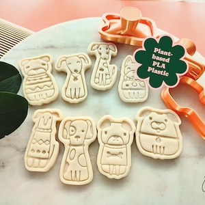 Puede incluir: Un conjunto de cortadores de galletas con forma de perro y galletas estampadas. Los cortadores son de color naranja y las galletas beige. Una pegatina verde con forma de flor dice "Plant-based PLA Plastic".