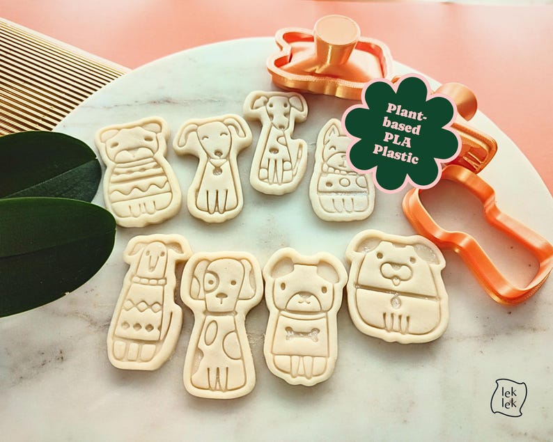 Puede incluir: Un conjunto de cortadores de galletas con forma de perro y galletas estampadas. Los cortadores son de color naranja y las galletas beige. Una pegatina verde con forma de flor dice "Plant-based PLA Plastic".