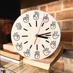 Puede incluir: Un reloj redondo blanco con gestos de manos negras que marcan las horas. El reloj tiene manecillas negras y un segundero rojo. Se exhibe en una estantería con libros y un fondo de pared de ladrillo.