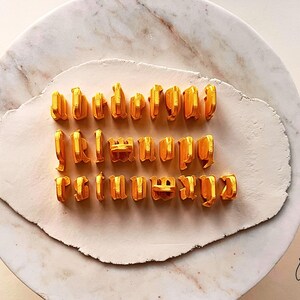 May include: A set of twenty-six individual golden yellow 3D-printed alphabet letter stamps arranged in three rows on a flat, off-white clay surface. Each letter features a distinct gothic-style typeface with a raised, textured structure.
