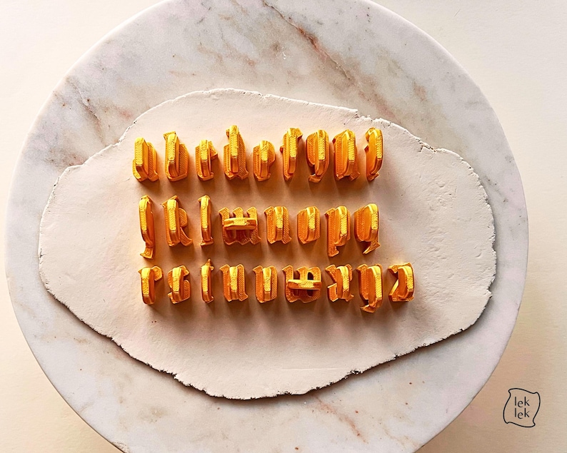 May include: A set of twenty-six individual golden yellow 3D-printed alphabet letter stamps arranged in three rows on a flat, off-white clay surface. Each letter features a distinct gothic-style typeface with a raised, textured structure.