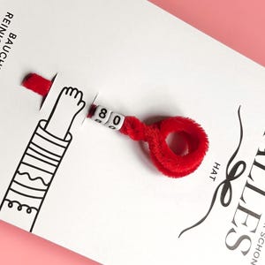 Peut inclure: Une carte blanche avec du texte allemand et un cure-pipe rouge flou attach&eacute;. Le cure-pipe porte le num&eacute;ro "80" sur de petites perles blanches. Un bras de dessin anim&eacute; est &eacute;galement sur la carte.