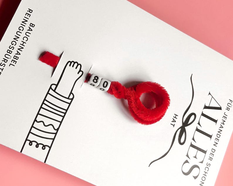 Peut inclure: Une carte blanche avec du texte allemand et un cure-pipe rouge flou attach&eacute;. Le cure-pipe porte le num&eacute;ro "80" sur de petites perles blanches. Un bras de dessin anim&eacute; est &eacute;galement sur la carte.