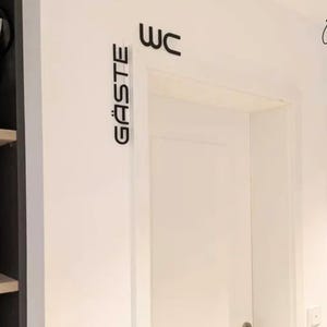 May include: A white door with black lettering that reads "WC" above the door and "G&Auml;STE" to the left. A small "lek lek" logo is in the upper right corner. The door is in a white room with a dark gray cabinet on the left.