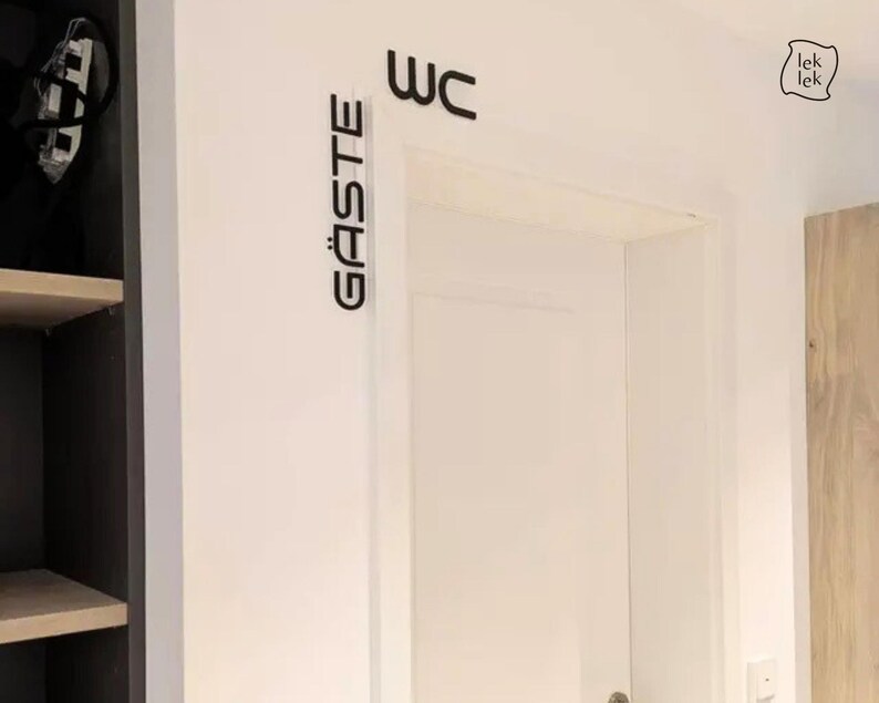 May include: A white door with black lettering that reads "WC" above the door and "G&Auml;STE" to the left. A small "lek lek" logo is in the upper right corner. The door is in a white room with a dark gray cabinet on the left.
