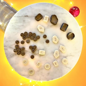 Op de afbeelding: Een verzameling bruine en witte plastic runen op een wit marmeren oppervlak. De runen zijn in verschillende vormen en maten, en sommige hebben symbolen die erin zijn gegraveerd.