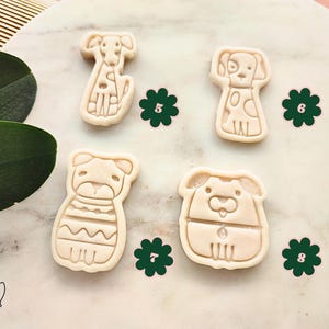 Puede incluir: Cuatro cortadores de galletas con forma de perro en varias poses. Los cortadores son de color beige y tienen contornos detallados. Decoraciones verdes en forma de flor se colocan alrededor de los cortadores. La imagen est&aacute; sobre una superficie similar al m&aacute;rmol.