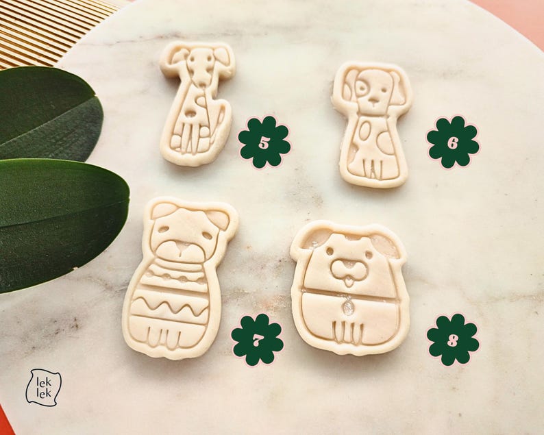 Puede incluir: Cuatro cortadores de galletas con forma de perro en varias poses. Los cortadores son de color beige y tienen contornos detallados. Decoraciones verdes en forma de flor se colocan alrededor de los cortadores. La imagen est&aacute; sobre una superficie similar al m&aacute;rmol.