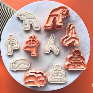 Pode incluir: Um conjunto de cortadores de biscoitos com poses de ioga, incluindo poses em pé, sentadas e de flexão. Os cortadores são de cor pêssego claro, com alguns em branco sujo contrastante. Estão dispostos sobre uma superfície branca com efeito de mármore.